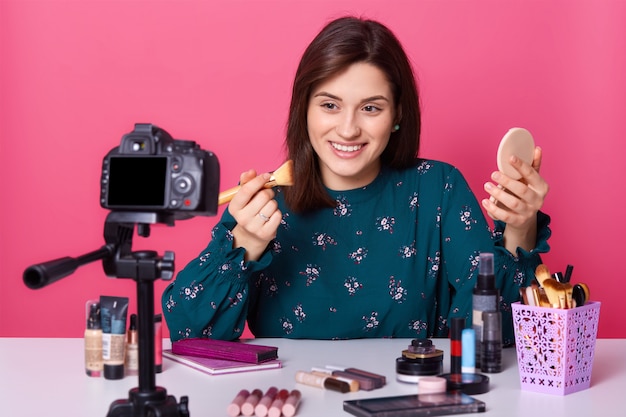 Sluit portret van het vrolijke schoonheid blogger stellen geïsoleerd over roze in studio, houdend professionele borstel voor omhoog maken