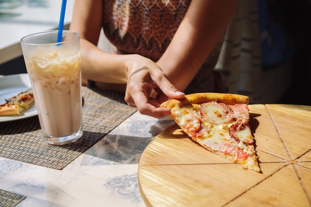 Sluiting van een onherkenbare vrouw die een heerlijke pizza vasthoudt met een glas koude koffie in zonlicht