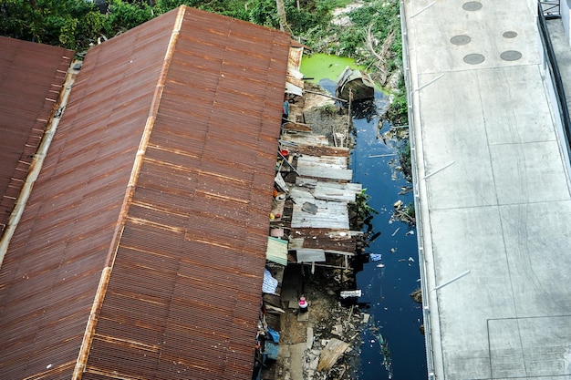 Photo slum zone, photo shooting from top view, can see rust rooftop, garbage and dirty water beside building.