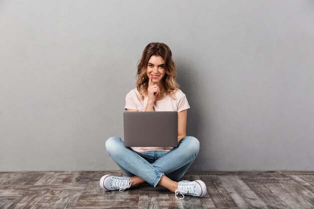 Sluwe glimlachende vrouw in t-shirt zittend op de vloer met laptopcomputer en over grijs