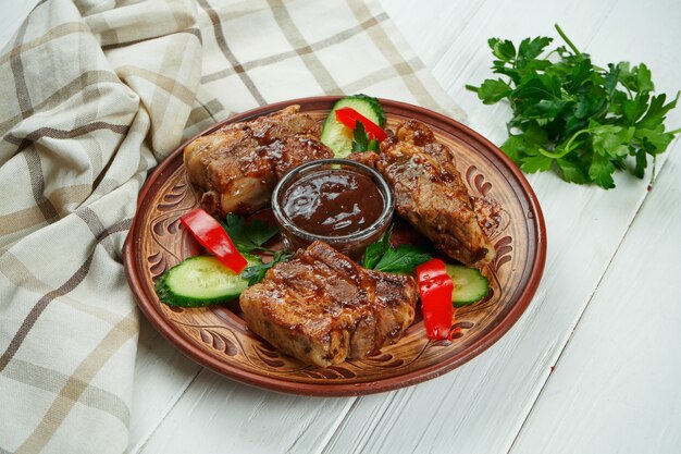 Smakelijk gebakken barbecue varkensribbetjes met rode saus en groenten in een keramische plaat op een witte houten achtergrond.