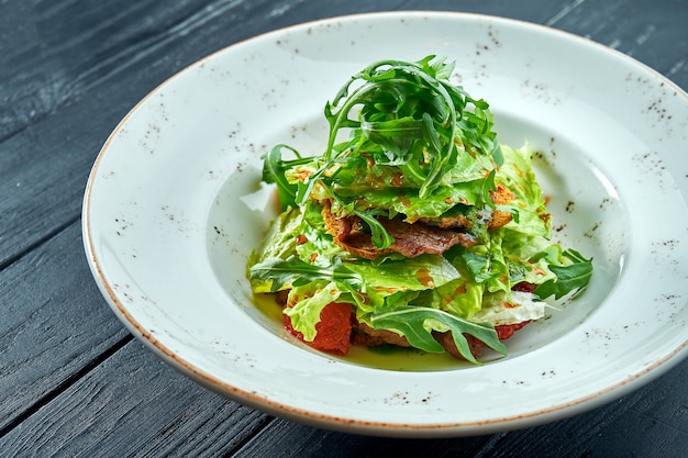 Smakelijke, dieetsalade met rucola, saus, sla en warm rundvlees in een witte plaat op donkere houten tafel