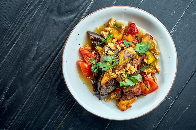 Smakelijke, dieetsalade van gebakken groenten (tomaten, paprika's, aubergines) met saus en noten in een witte plaat op een donkere houten ondergrond. Vegetarisch eten. Selectieve focus