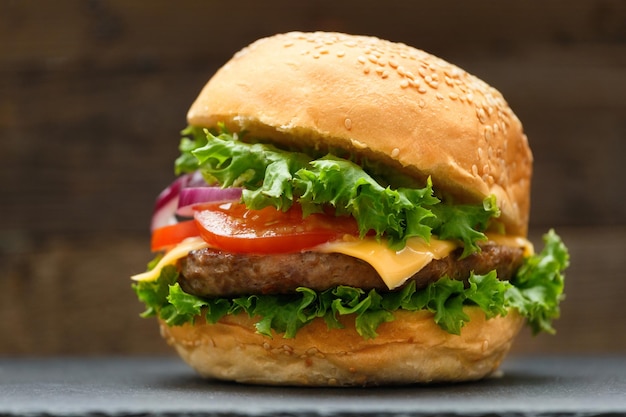 Smakelijke hamburger met sappige rundvleeskotelet, kaas, tomaten, sla, ui op een zwarte stenen plaat op een donkere houten achtergrond. Fastfood zelfgemaakte hamburger