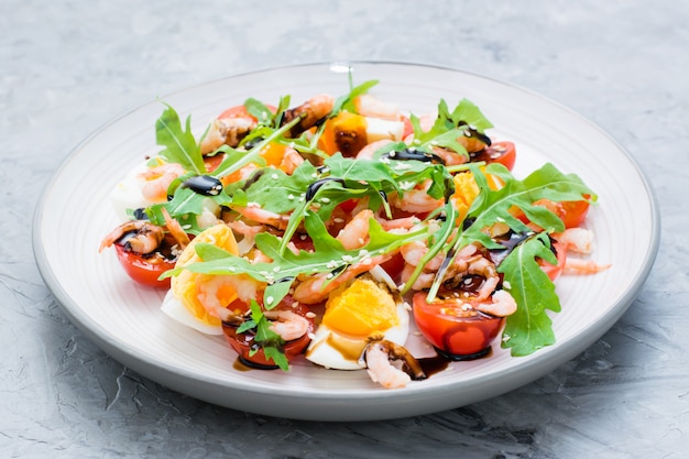 Smakelijke salade van kerstomaatjes, eieren, gekookte garnalen, rucola, sesamzaad en balsamico-saus in een bord