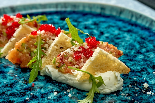 Smakelijke salade - zalmtartaar met witte espuma, rucola, croutons en tobiko-kaviaar in een blauw bord op een lichtblauwe achtergrond