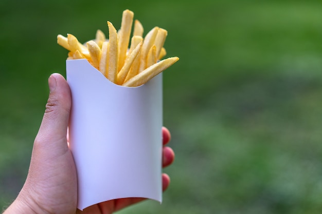 Smakelijke sappige frietjes in witboekvakje in mensenhand op aard in openlucht. fast-food mockup ongreen achtergrond. ongezond voedselconcept met vrije ruimte voor tekst. gebakken aardappelmenu.