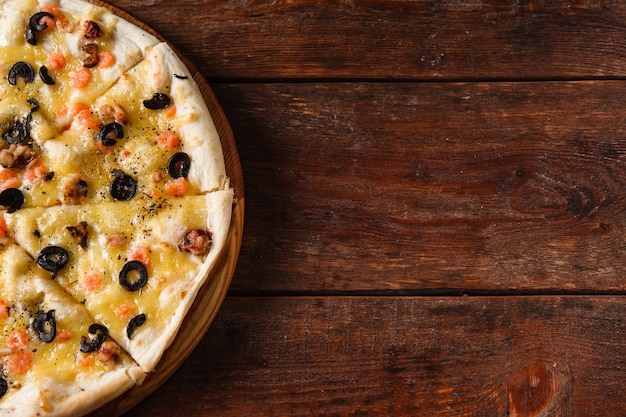 Smakelijke verse Italiaanse pizza met garnalen, olijven en kaas geserveerd op rustieke houten tafel. Donkere achtergrond met vrije ruimte voor tekst.