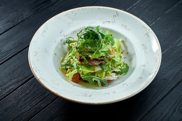 Smakelijke, voedingssalade met rucola, saus, sla en warm rundvlees in een witte plaat op een donkere houten achtergrond.