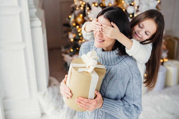 Small adorable female kid prepares surprise for her mother closes eyes and gives wrapped present as stand at New Year background Pretty female recieves gift from daughter Surprisment concept