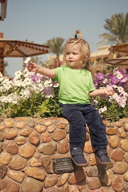 small baby boy or cute child with adorable face and blonde hair in green shirt and pants sitting on stony parapet playing with flowers sunny outdoor on natural blue sky background