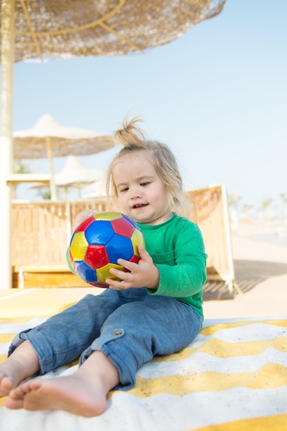 Small baby boy or cute child with happy face and blonde hair in green shirt and pants sitting barefoot playing with colorful ball at sunny summer outdoor beach on natural sky background