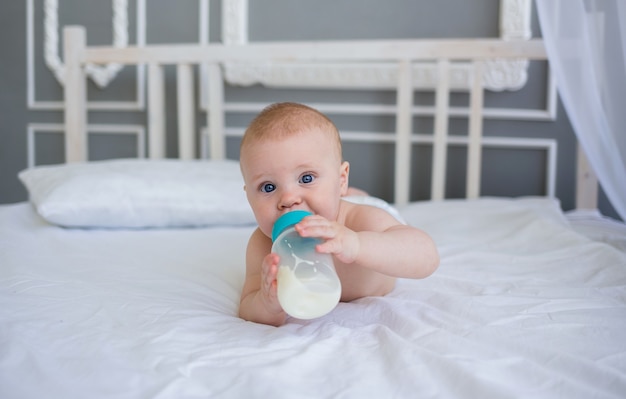 Small baby in a diaper is lying on the bed and drinking milk from a bottle
