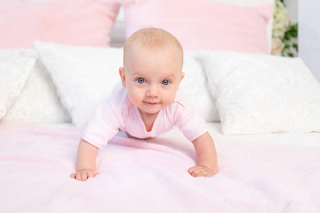 Piccola neonata di 6 mesi che striscia su un letto bianco e rosa a casa, guardando la telecamera, posto per il testo