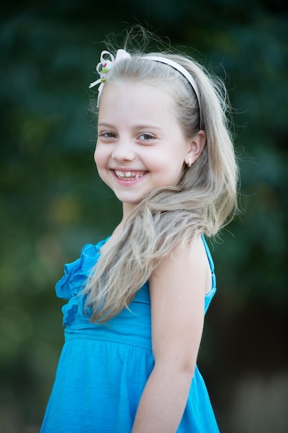 작은 아기 소녀 또는 귀여운 행복한 아이가 사랑스러운 웃는 얼굴을 하고 여름에 파란 드레스를 입고 흐릿한 배경에서 야외에서 금발 머리에 절을 합니다.