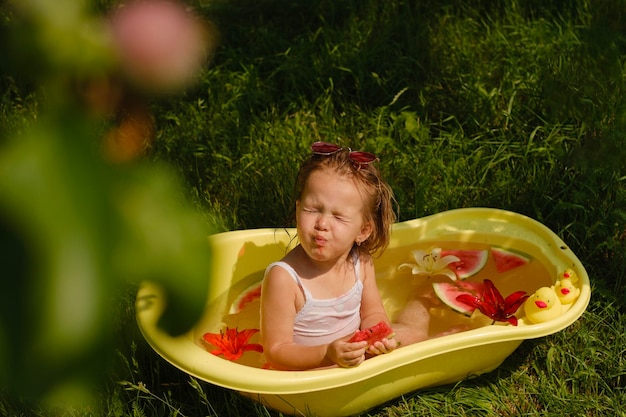 Small baby splashes in the garden in the bathtub and eats a watermelon