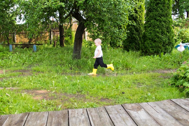 A small baldheaded boy in yellow boots runs in the countryside through puddles in the fresh air