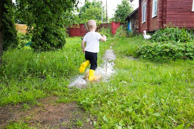 黄色いブーツをはいた禿頭の小さな男の子が、新鮮な空気の中で水たまりを通り抜けて田園地帯を走る