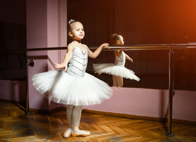 Small ballerina girl in white tutu standing next to the mirror with her reflection in it at dance studio Children and ballet concept