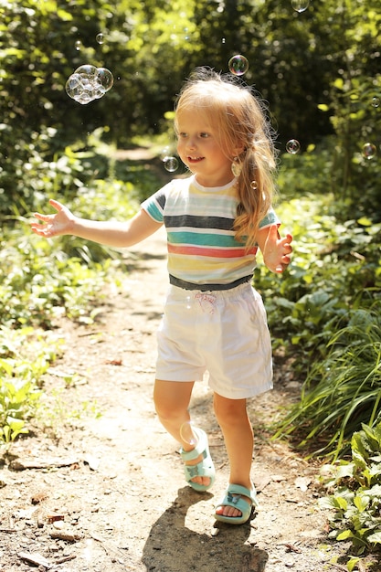 軽いTシャツと白いパンツを着た小さな美しいブロンドの女の子が公園でシャボン玉をキャッチ