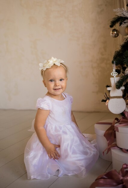 Photo a small beautiful girl in a festive white dress sits near the christmas tree
