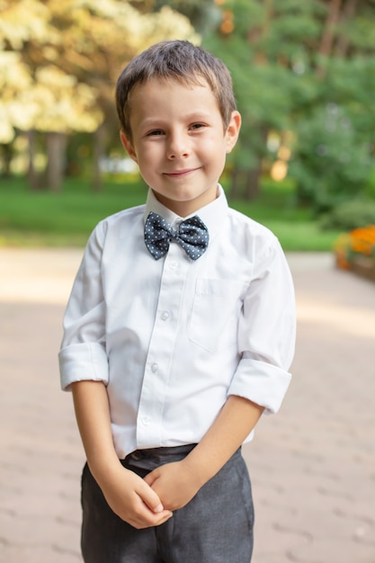 a small beautiful schoolboy boy in a white shirt and gray trousers is standing on the street