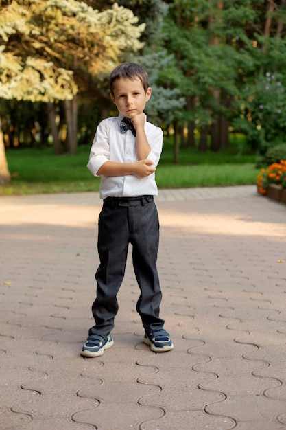 a small beautiful schoolboy boy in a white shirt and gray trousers is standing on the street