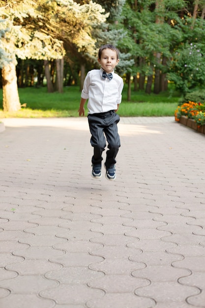 a small beautiful schoolboy boy in a white shirt and gray trousers jumps on the street