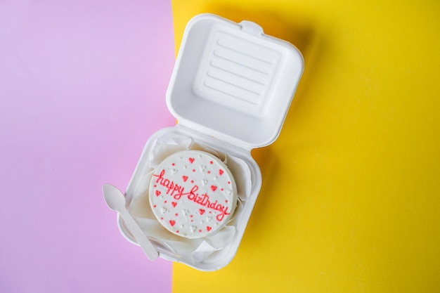 A small bento birthday cake in a box Wooden spoon with white ribbon Yellow and pink background