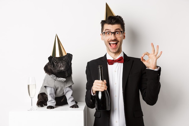 Small black dog wearing party hat and standing near happy man celebrating holiday, owner showing okay sign and holding champagne bottle, white background.