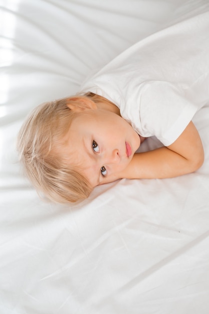 small blond boy in a white Tshirt is lying on his stomach on a white sheet in the bed