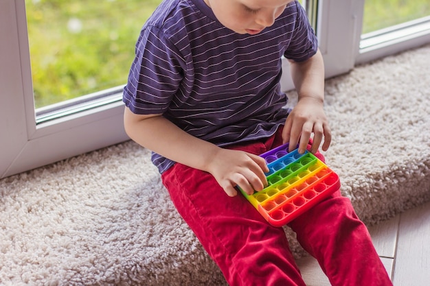 小さな金髪の少年現代のカラフルで明るいポピットのおもちゃで遊んでいる小さな金髪の少年シンプルなディンプル。カラフルな流行の抗ストレスシリコン感覚玩具を持つ子供