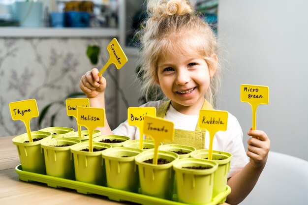 エプロンを着た小さなブロンドの女の子は、子供たちのガーデニングの概念である笑顔で、苗の種を植えることに従事しています。