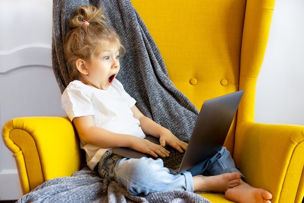 작은 금발 소녀는 매우 놀라고 겁에 질린 표정으로 앉아 노트북을 봅니다.