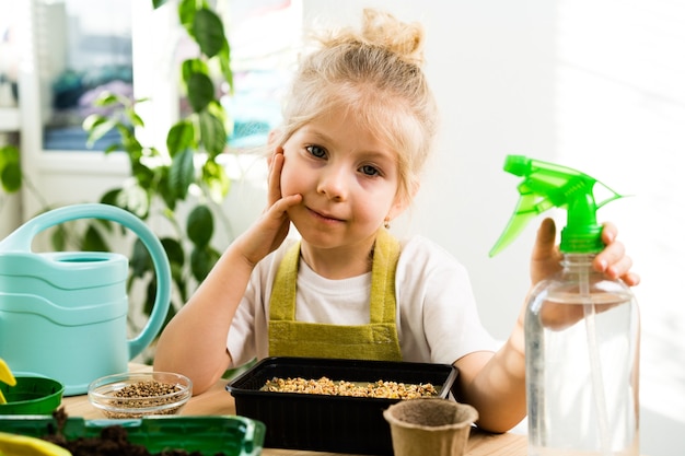 A small blonde girl sits brooding at a wooden table with her cheek propped on her hand, growing micro greens, watering and spraying with water from a pulevizer.