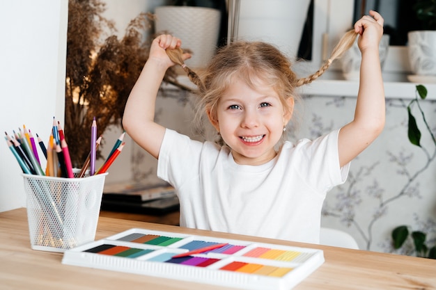 A small blonde girl with a charming smile sits at a table with a bright colorful palette