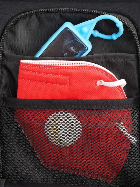 Маленькая бутылочка с дезинфицирующим средством для рук с красной защитной маской в кармане черного рюкзака.
