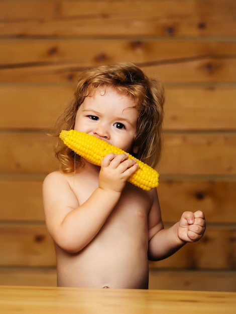 Small boy child with long blonde hair eating yellow corn or maize with bare chest and funny face on wooden or wood background