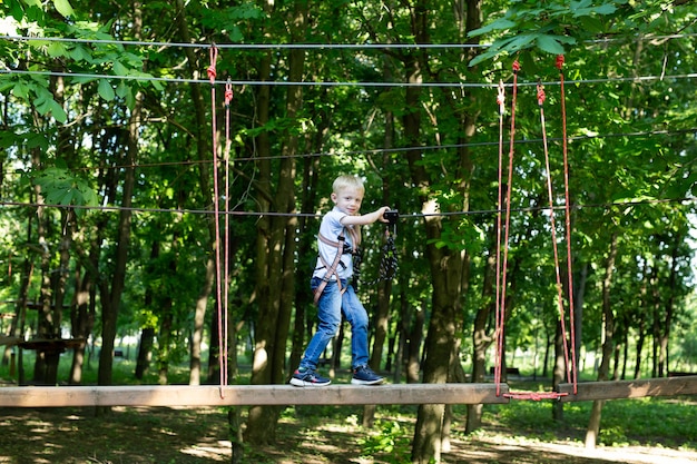 Un bambino in attrezzatura da arrampicata sta camminando lungo una strada di corda in un parco avventura, aggrappandosi a una corda e un moschettone.