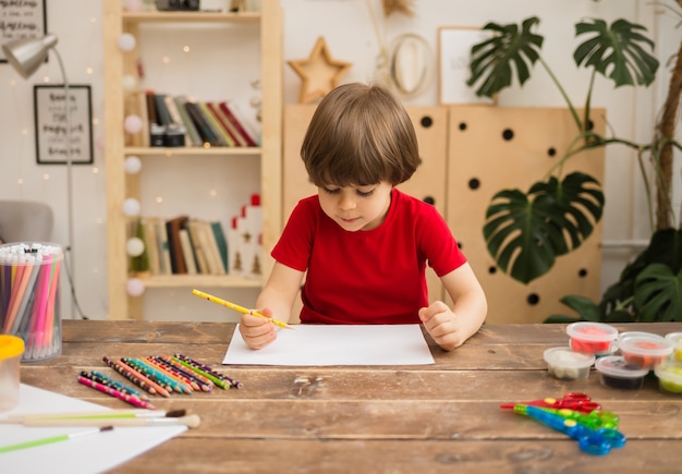 Small boy draws with a pencil on white paper at a wooden table with stationery