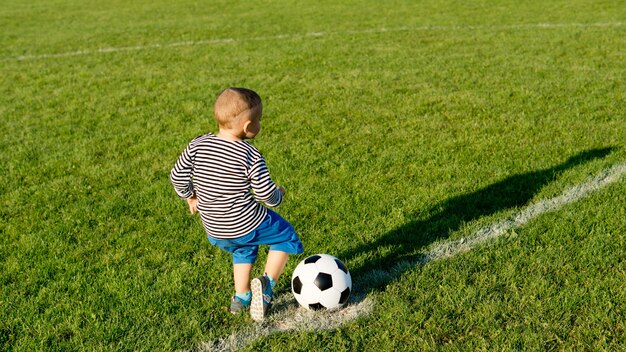 夕方の光の中で彼のサッカーボールを蹴って緑のスポーツフィールドを横切って走って楽しんでいる小さな男の子