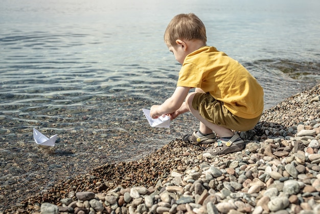 Small boy is launching white paper boats into the clear water of a large lake with a stony bottom