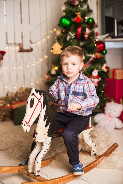 Small boy riding wooden rocking horse in front of christmas tree