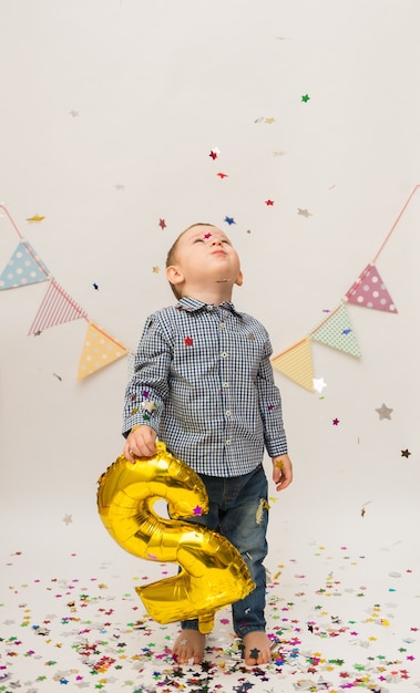 Small boy in a shirt and trousers stands and holds a gold foil number two