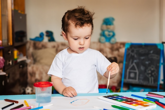 A small boy sits on a chair and draws with colored paints