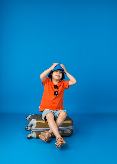 Small boy in a T-shirt and shorts and a panama hat sits on a suitcase on a blue surface with space for text