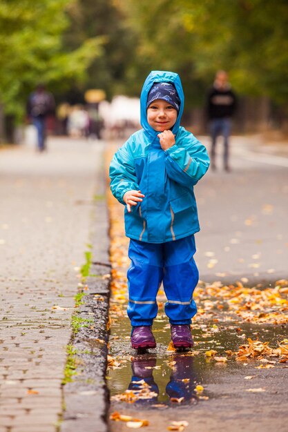 防水服を着た小さな男の子がアスファルト道路の水たまりに立っています