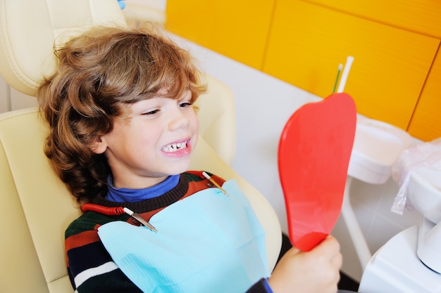 A small boy with curly hair in a dental chair opening his mouth to show where he lost one of his baby's teeth