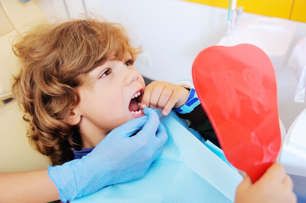 A small boy with curly hair in a dental chair opening his mouth to show where he lost one of his baby's teeth