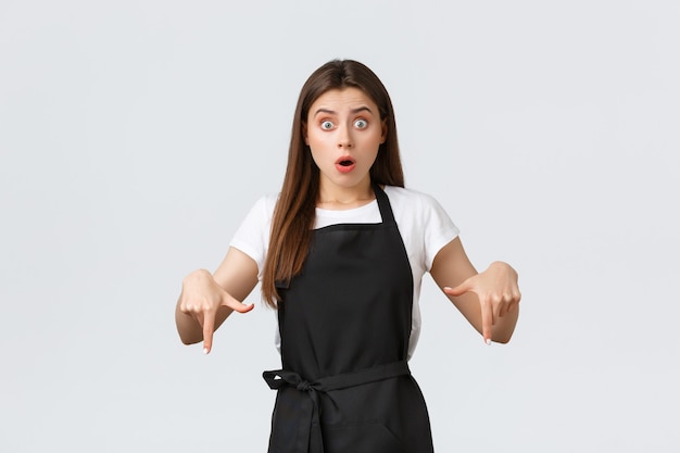 Small business, employees and coffee shop concept. Concerned and shocked waitress pointing fingers down. Gasping confused barista asking question. Astonished saleswoman tell big news